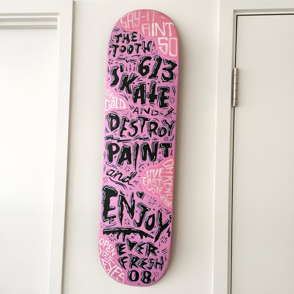 A vertical pink skateboard featuring bold graffiti-style artwork is mounted on a white wall, showcasing creative skateboard wall art with an edgy, urban aesthetic for a girls bedroom.