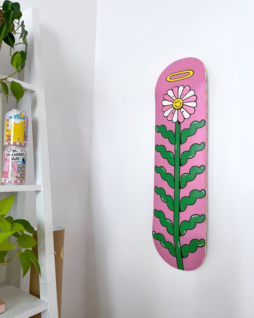 A pink skateboard wall art piece featuring a whimsical flower with a smiling face and halo is mounted on a white corner wall, adding a playful and artistic touch to a girl's bedroom.