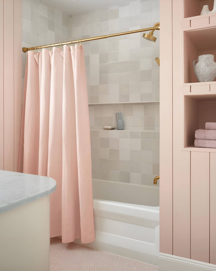Soft and elegant girls bathroom design featuring blush pink accents, a gold shower fixture, and built-in shelving with neatly folded towels and decor.