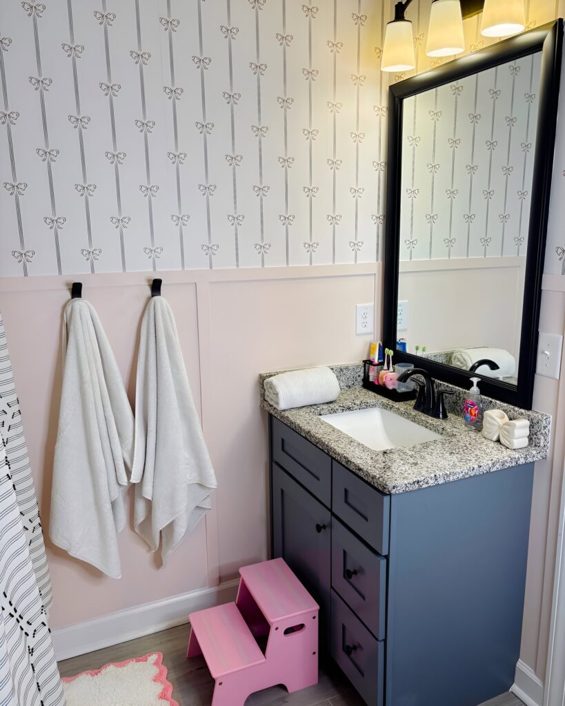 Stylish and functional bathroom design for girls featuring a granite vanity, bow-patterned wallpaper, soft pastel colors, and a pink step stool for easy sink access.