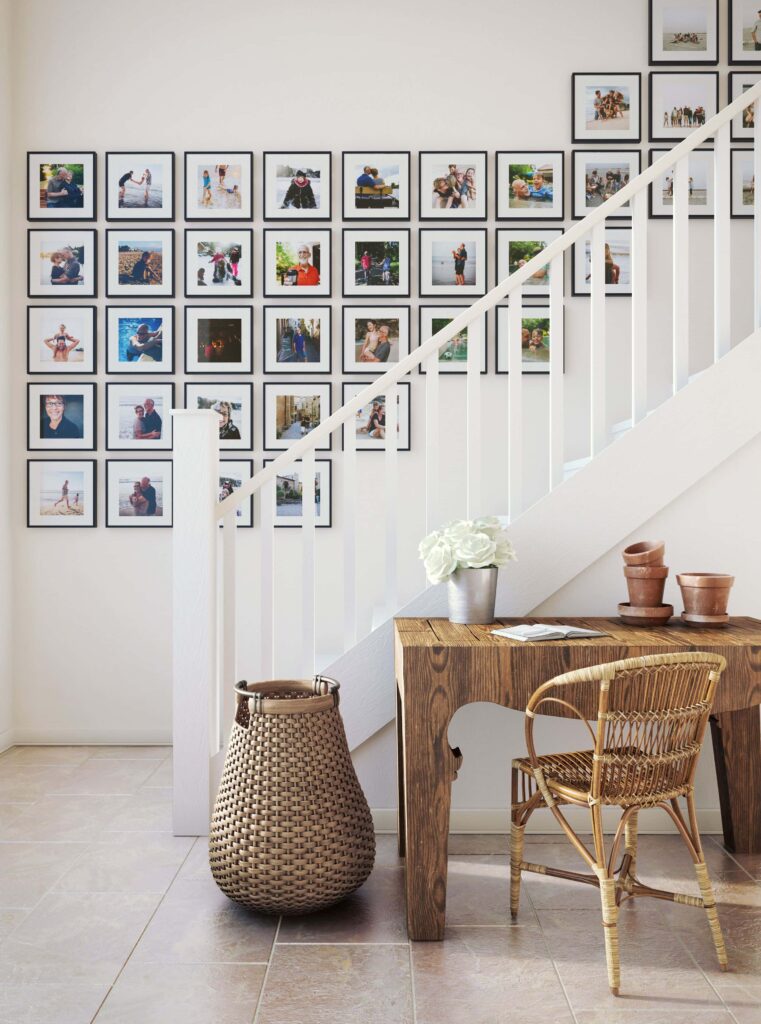A bright and modern staircase memory wall displaying a symmetrical grid of colorful family photos in black frames alongside a rustic wooden desk and wicker chair.