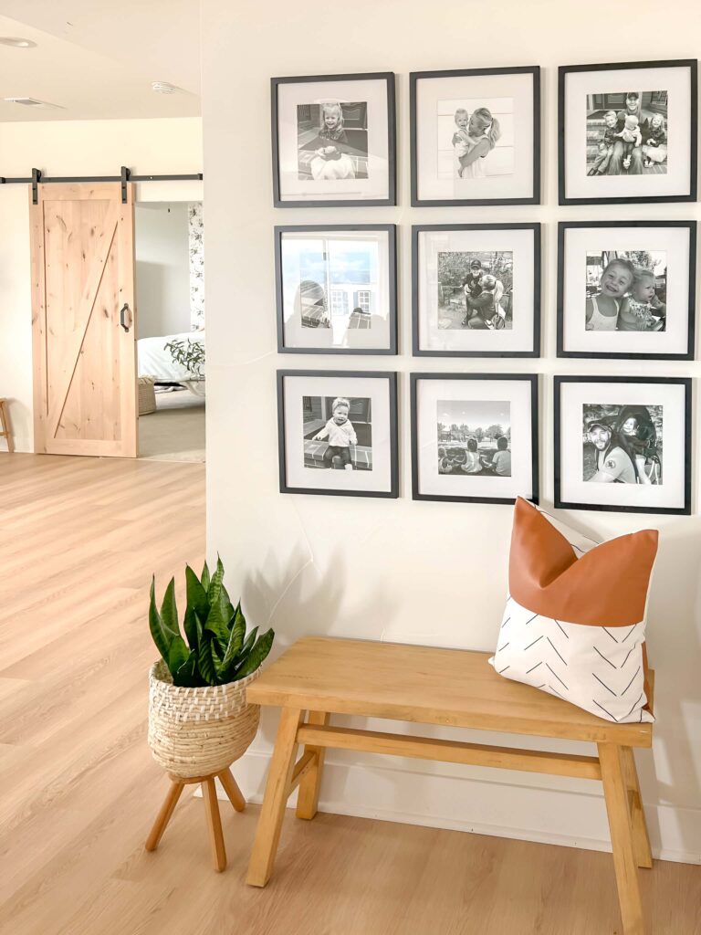 A cozy corner featuring a family memory gallery wall with nine black-and-white framed photos arranged in a grid above a wooden bench adorned with a stylish pillow and a potted plant nearby.