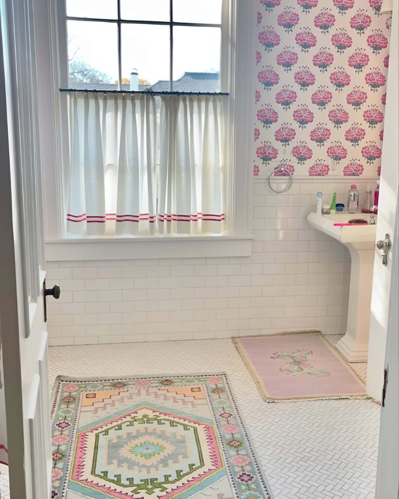 Charming girls' bathroom design featuring pink floral wallpaper, playful pastel rugs, and a vintage-style sink with white tile walls and floors.