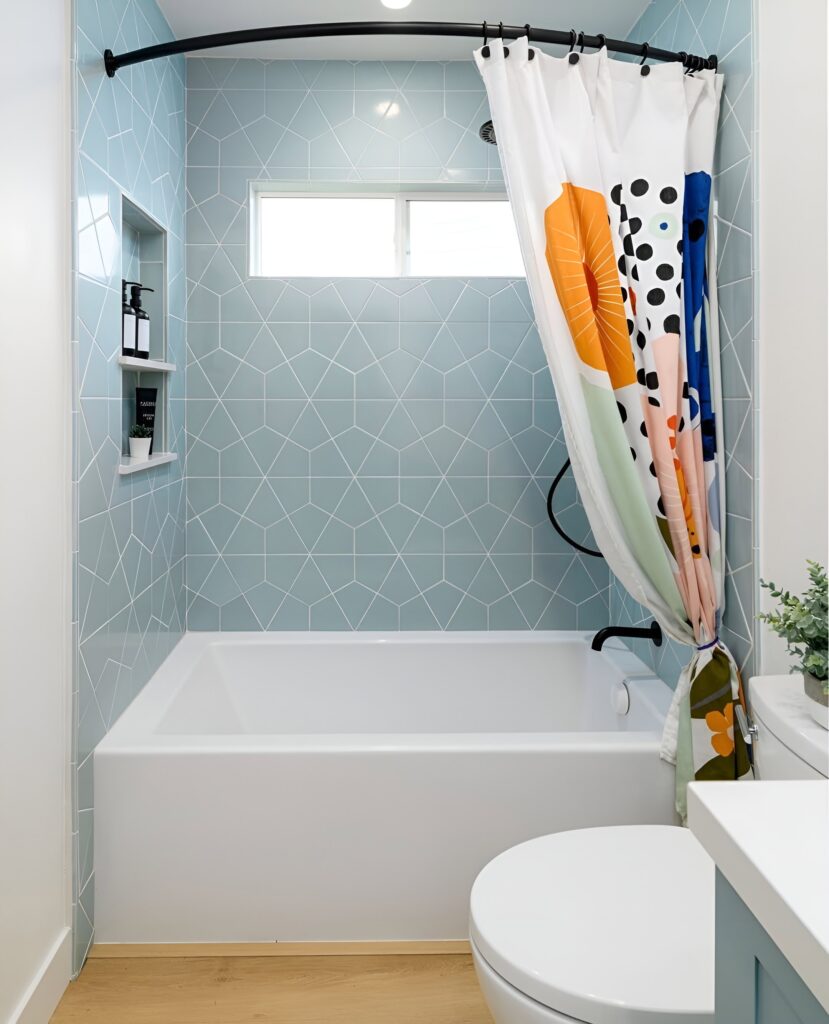 A modern kids bathroom design features light blue geometric wall tiles, a white bathtub, a colorful patterned shower curtain, and wooden flooring.