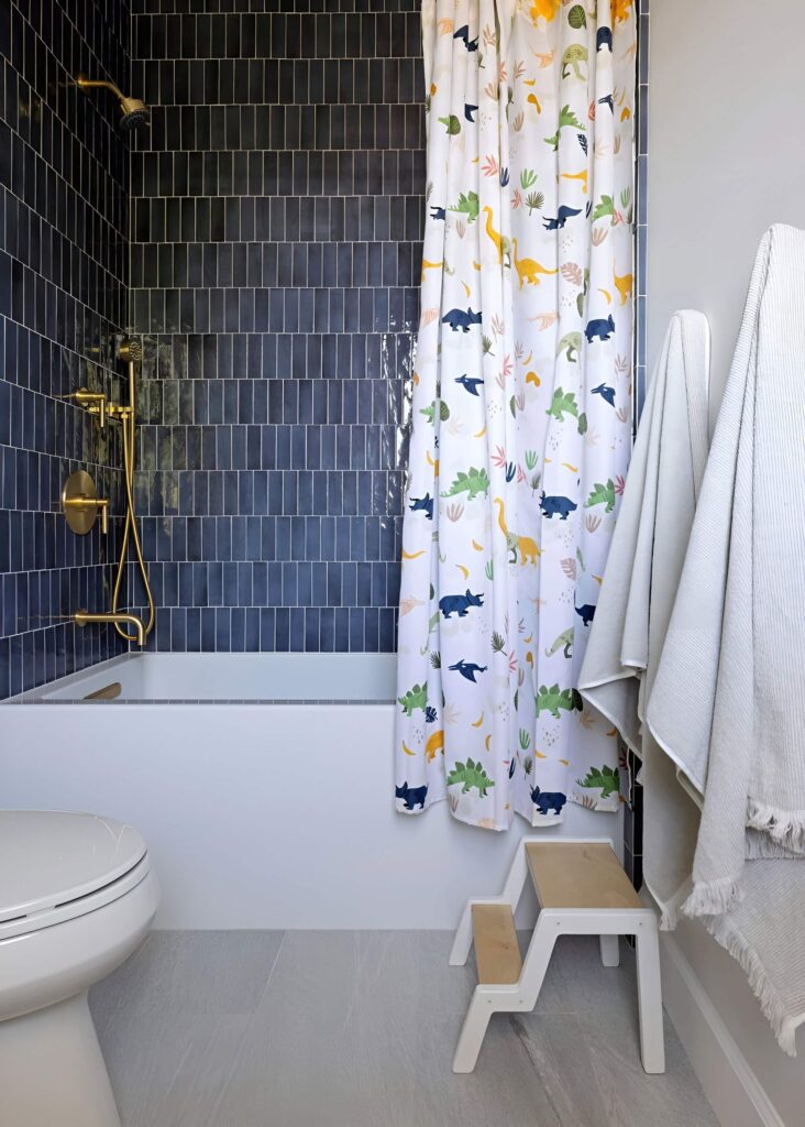 A modern bathroom featuring a dinosaur-themed shower curtain, sleek navy tiles, and a wooden step stool, providing creative bathroom design ideas for a boy’s space.