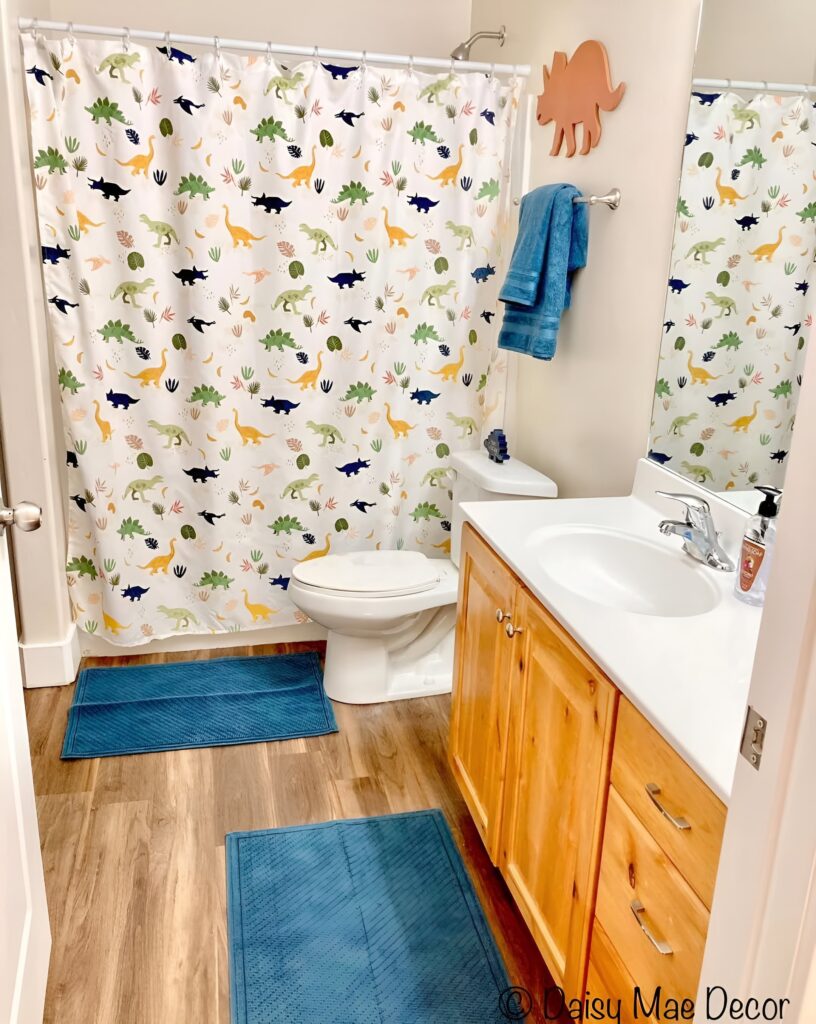A fun and vibrant bathroom with a dinosaur-themed shower curtain, wooden vanity, and blue bath mats, offering playful bathroom design ideas for a boy's space.