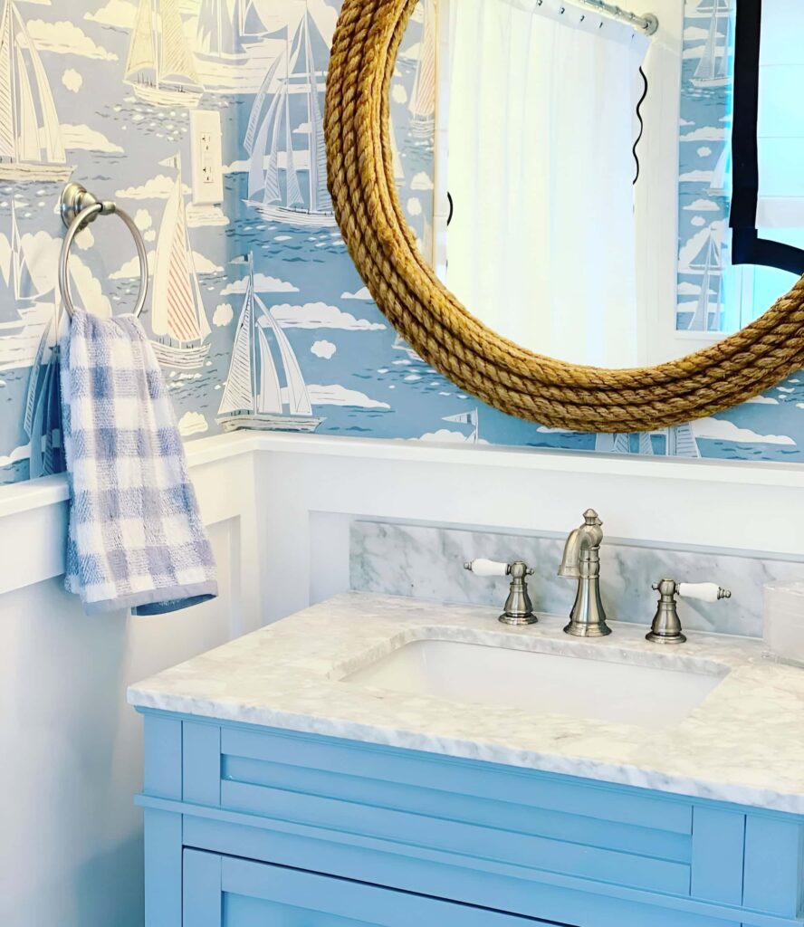 A playful and nautical-inspired bathroom design with blue tones, featuring a marble countertop, a rope-framed mirror, and sailboat-themed wallpaper, offering bathroom design inspiration for a kid's space.