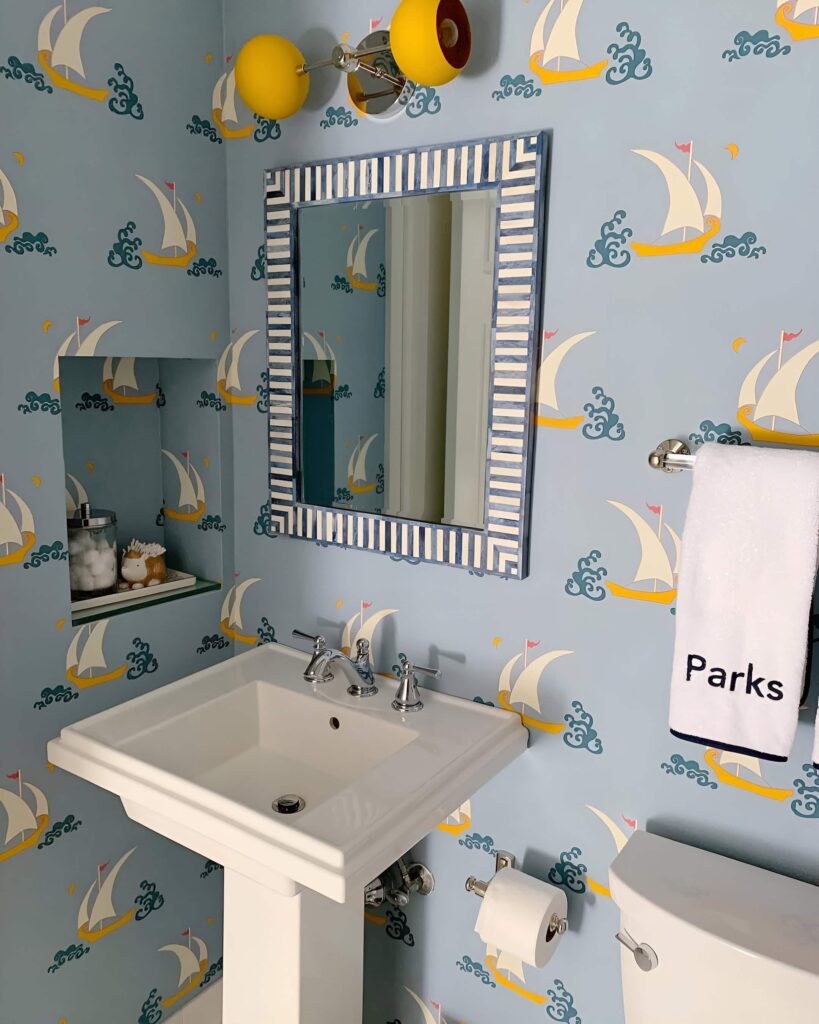 A bright and cheerful bathroom with playful sailboat wallpaper, a striped mirror, and colorful lighting, offering bathroom design inspiration for a kid-friendly space.