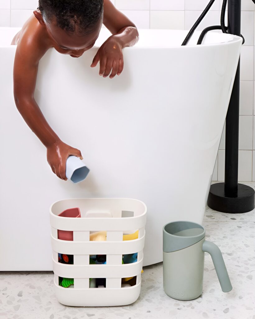 A young boy reaches out of a modern white bathtub to place a toy in a stylish storage basket, showcasing practical bathroom design for kids.