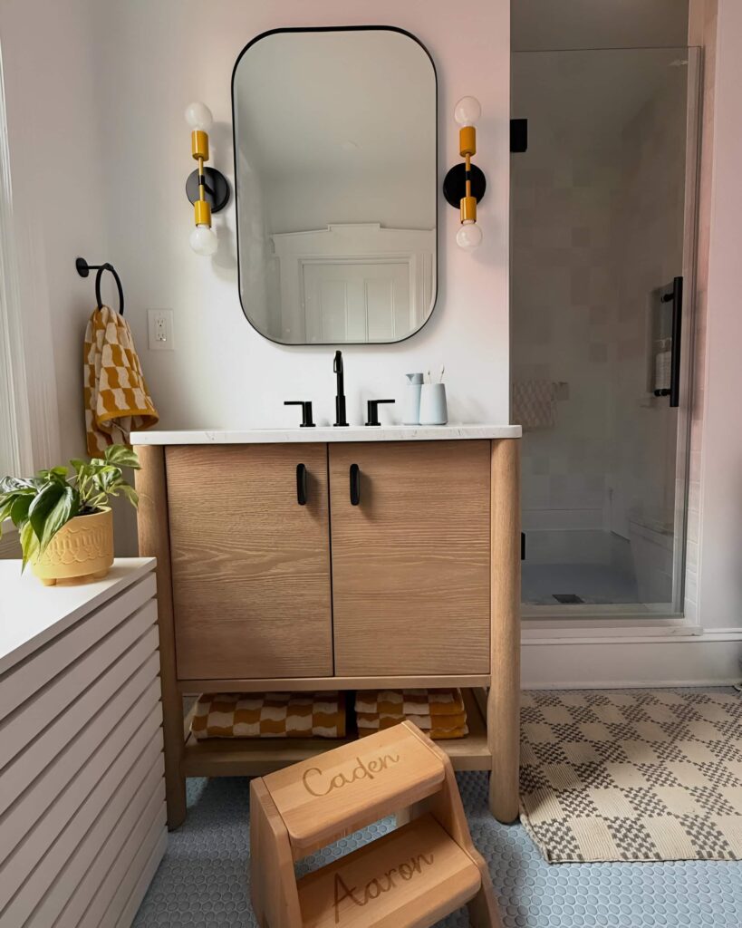 Modern kids’ bathroom design featuring a wood vanity with black hardware, double light sconces, a step stool, and a walk-in shower.