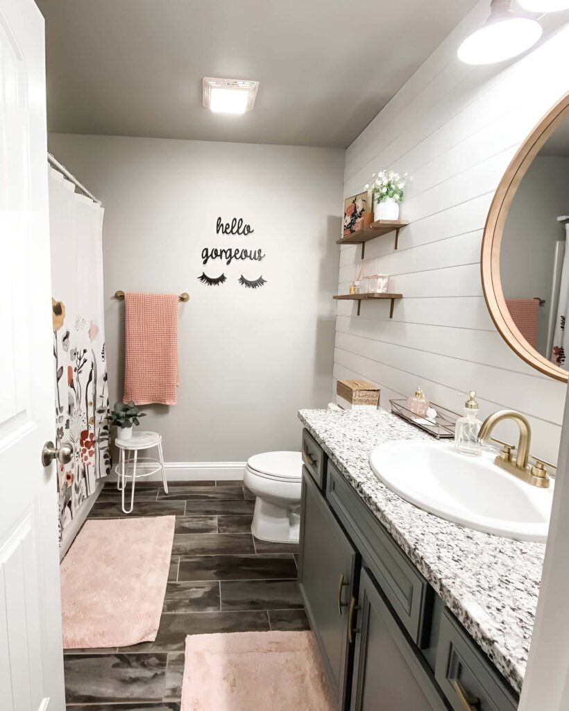 Cozy and stylish girls bathroom design featuring soft pink accents, floral shower curtain, wood shelves, and a round mirror over a granite countertop vanity.
