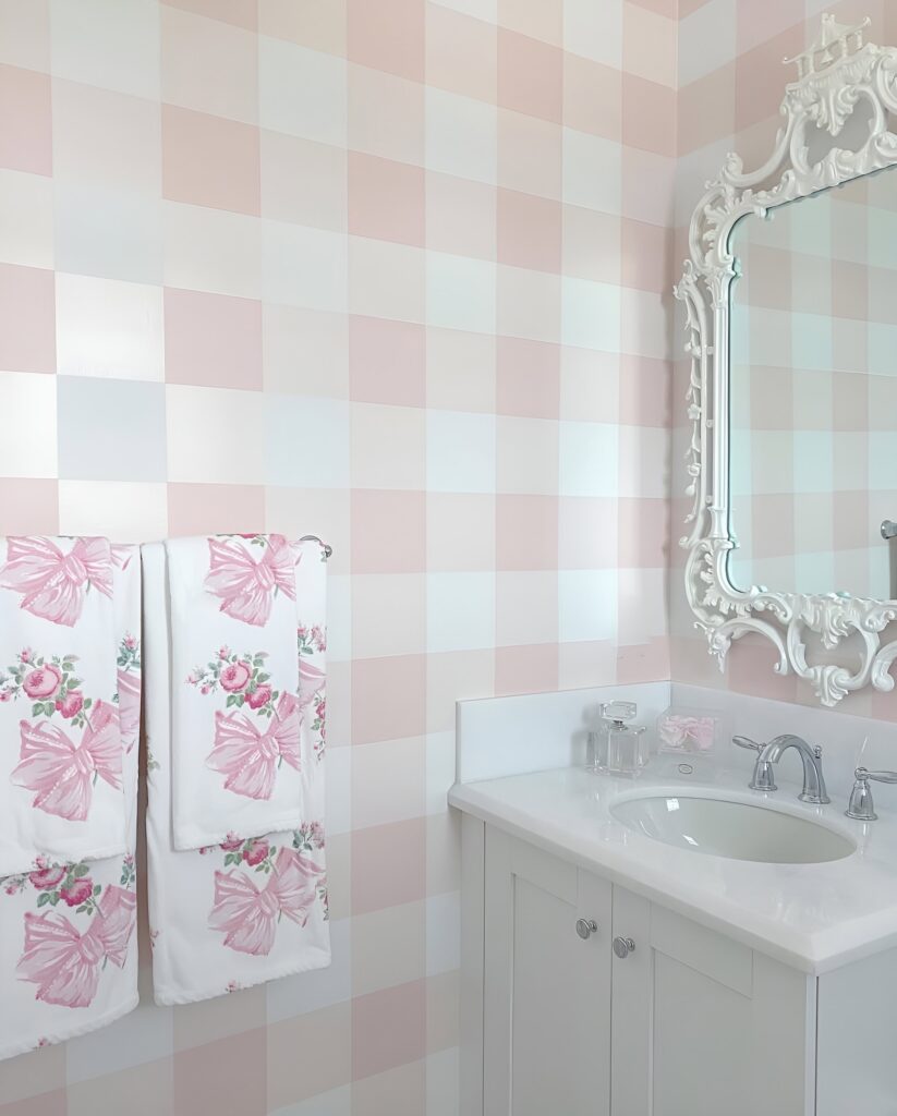 Sweet and feminine girls' bathroom design with pink gingham wallpaper, floral bow towels, a white vanity, and an ornate vintage-style mirror.