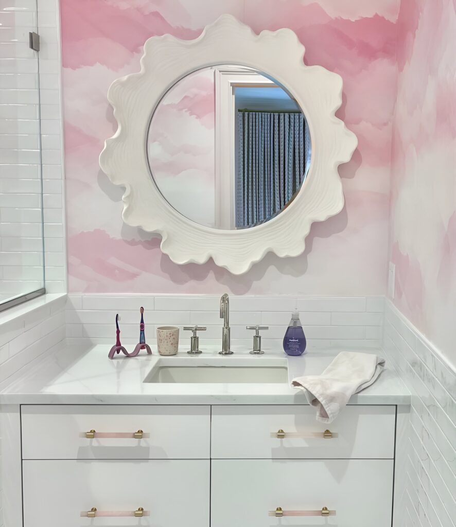 Whimsical girls' bathroom design with pink cloud-patterned wallpaper, a scalloped round mirror, and a white vanity with gold and pink drawer pulls.