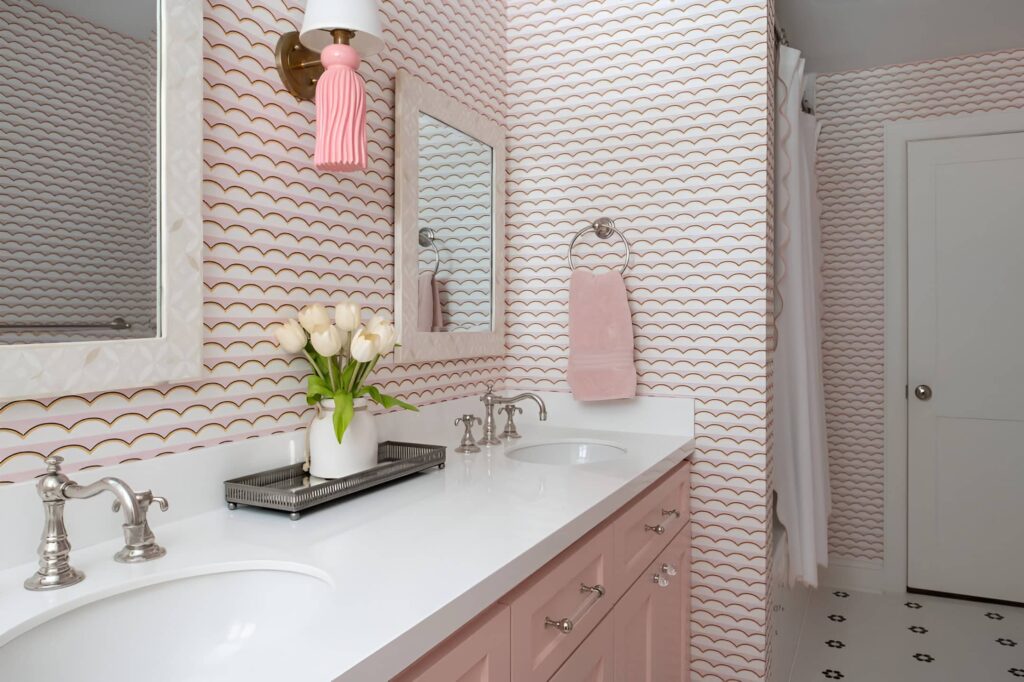 Playful girls' bathroom design with pink scalloped wallpaper, double sinks, soft pink cabinetry, and floral decor for a cheerful and feminine touch.