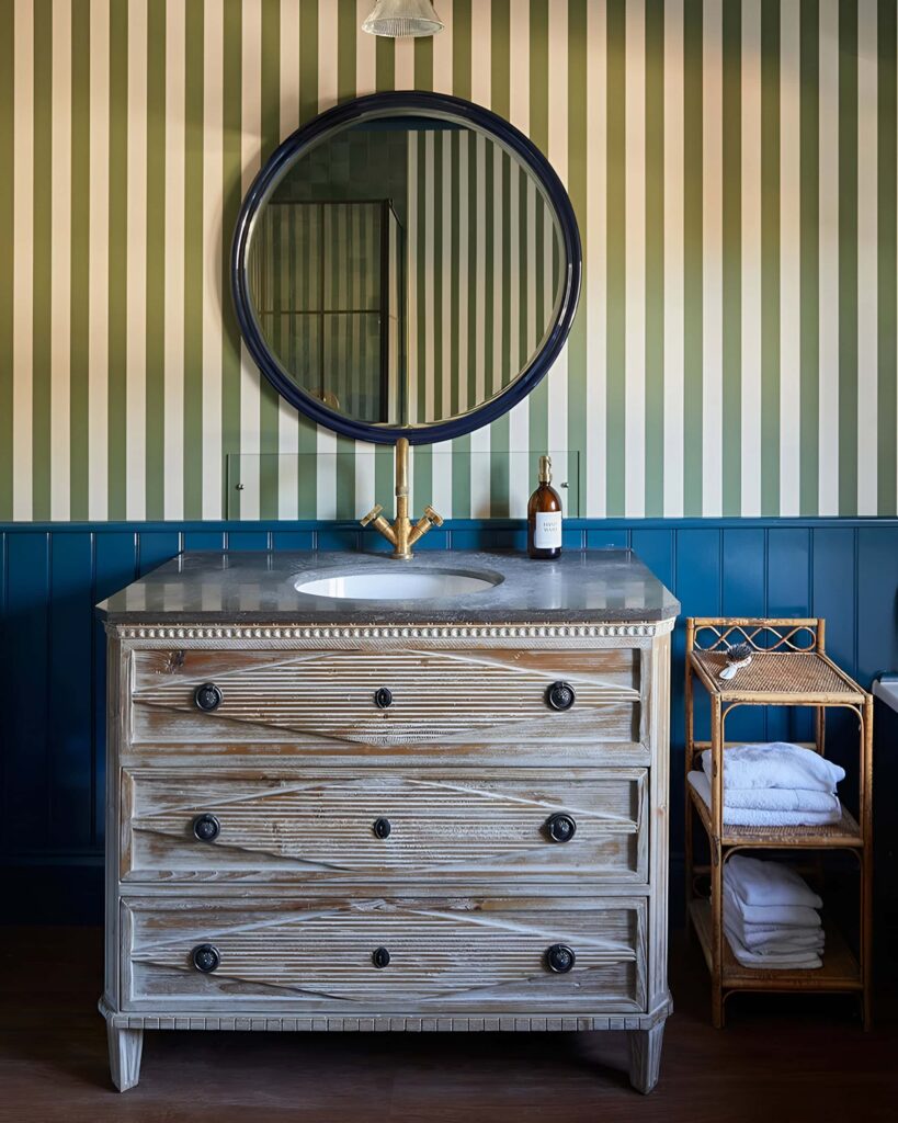 Vintage-inspired kids’ bathroom design featuring a striped wallpaper, distressed wood vanity with a marble top, round mirror, and gold faucet.